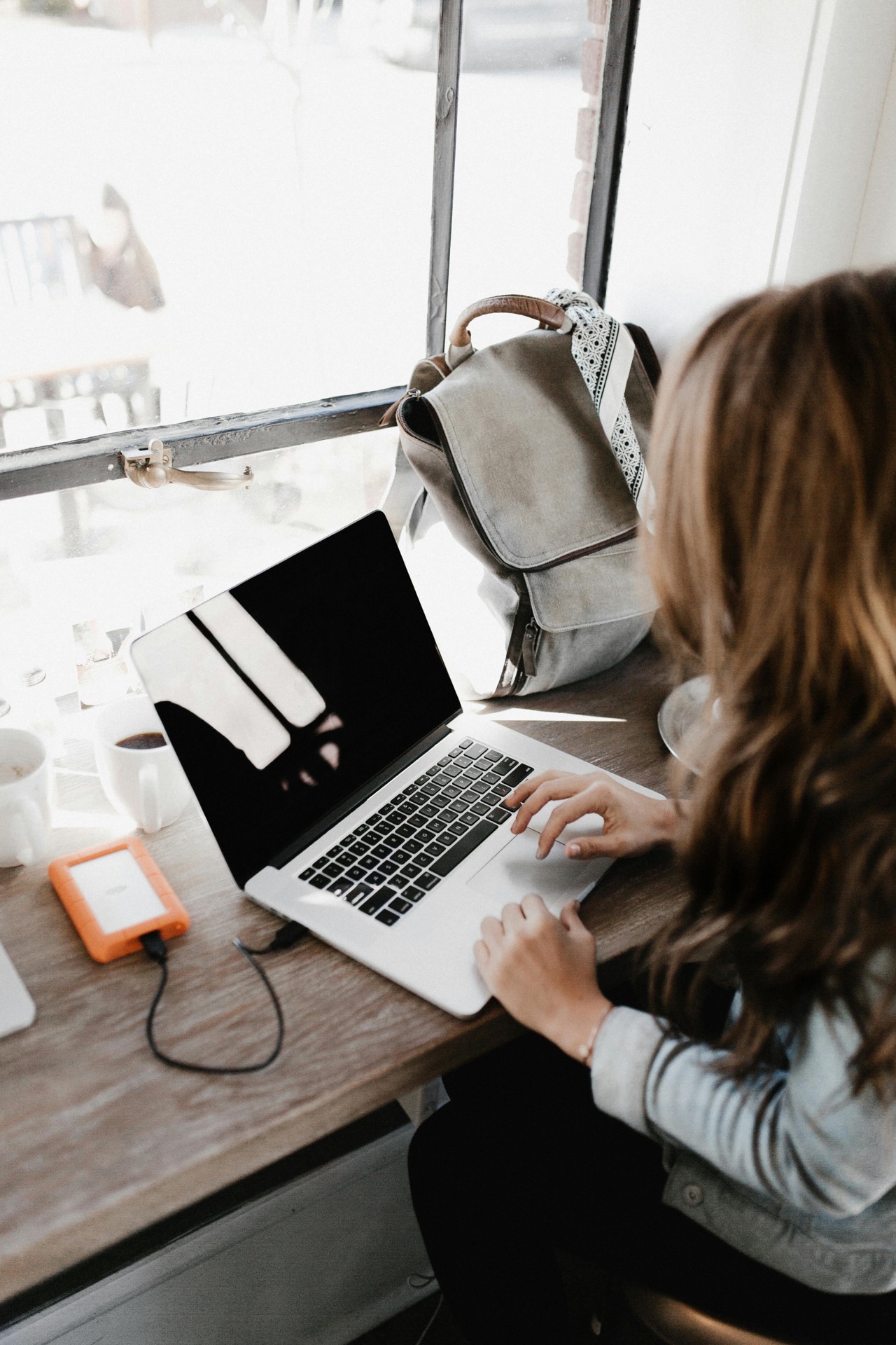 Meisje met lichtbruin haar zit aan een verhoogde tafel op haar laptop te werken. Haar telefoon ligt op te laden aan haar laptop en naast haar staat haar tas en een kopje koffie.